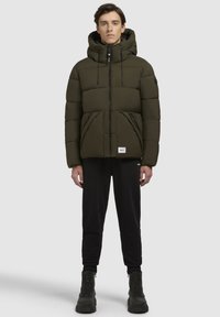 Chaqueta acolchada verde olivo con capucha, que presenta un cierre frontal con cremallera, paneles cosidos y bolsillos laterales. Lleva puesta con pantalones de chándal negros y botas.