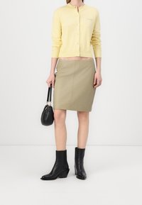Yellow knitted cardigan with round neckline, button closure, and light texture. Olive green pencil skirt, knee-length. Black ankle boots and handbag.