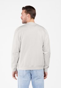 Hombre de espaldas vistiendo un sudadera beige clara y jeans azules claros frente a un fondo blanco liso.