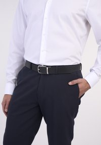 Schwarzer, strukturierter Gürtel mit silberner Schnalle, getragen auf der Hüfte über dunklen Hosen und einem weißen Hemd mit Knopfleiste, das einen formellen Stil präsentiert.