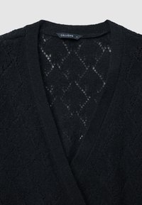 Cardigan nero lavorato a maglia con design a V. Presenta un motivo a rombi e una finitura testurizzata. L'etichetta riporta "CALLIOPE" in un colore a contrasto.