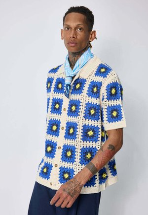 Personne portant une chemise en crochet bleu et blanc à motifs carrés, foulard bleu noué autour du cou, et pantalon bleu marine, debout devant un fond uni.