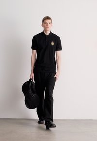 Melna polo krekls ar izšūtu emblēmu, kopā ar melnām biksēm. Modelis nēsā apaļu melnu sporta somu. Gluda auduma, vienkāršs dizains.