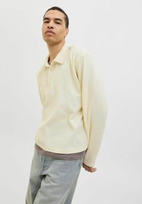 Polo shirt crème texturé à manches longues avec un col, doté d'une patte de boutonnage à trois boutons, porté par-dessus une couche grise, associé à un jean bleu clair.