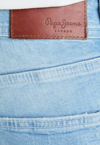 Heledad sinised denim püksid, millel on pruun nahkne tagakotike, millel on mulje "Pepe Jeans London", esitlevad õmblustead ja taskuõmblust.