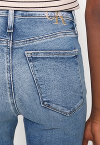 Los vaqueros de denim azul claro presentan un bolsillo trasero con un detalle de costura en la parte superior y un sutil logo dorado "CK" encima de la cinturilla.