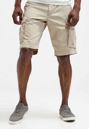 Khaki Cargo-Shorts mit Seitentaschen aus leichtem Stoff. Sie haben eine lockere Passform, gesäumte Kanten und ein schlichtes Design. Kombiniert mit grauen Turnschuhen.