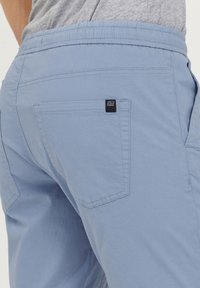 Pantaloni scurți de culoare bleu deschis, confecați dintr-un material moale, cu un tiv elastic. Au două buzunare în spate și o mică etichetă logo de culoare bleumarin.