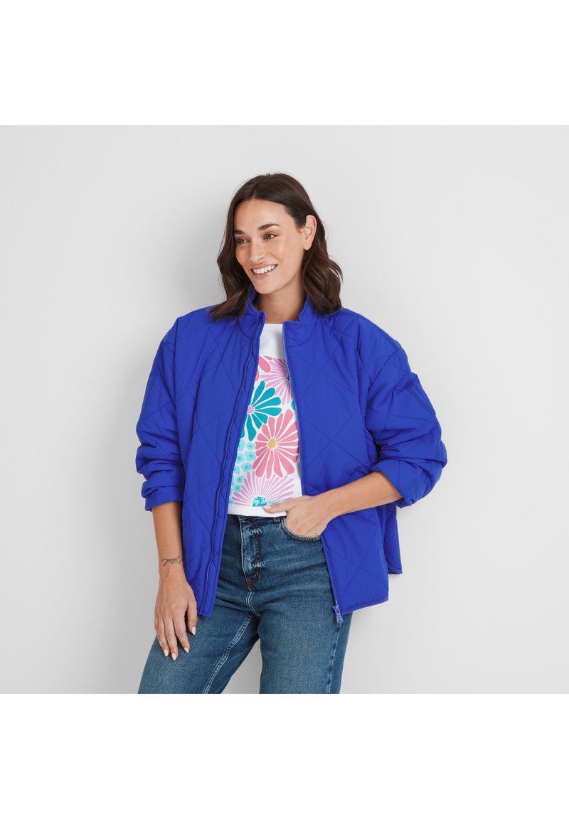 Woman with dark hair wearing blue quilted jacket, floral print white T-shirt, and blue jeans, smiling and posing with hand in pocket.