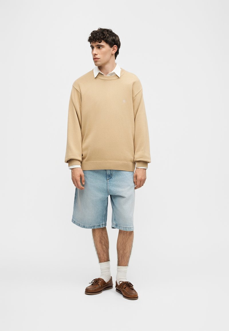 Jeune homme debout portant un pull beige sur une chemise blanche, un short en jean bleu clair, des chaussettes blanches et des chaussures bateau en cuir marron.