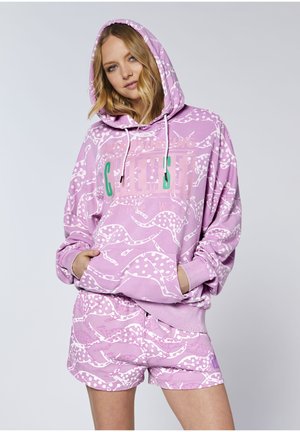 Junge Frau trägt einen pinken Kapuzenpullover und passende Shorts mit weißen Gepardenmustern, steht mit den Händen in den Taschen.