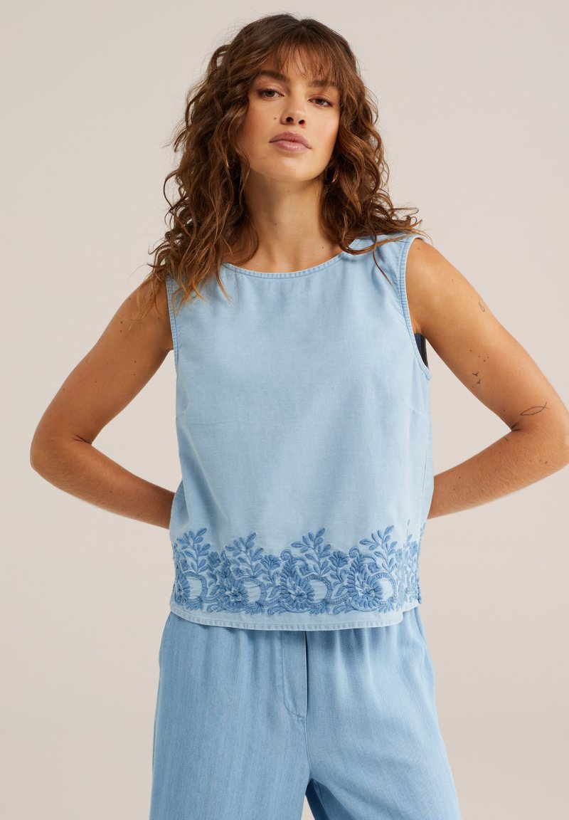 Femme aux cheveux bouclés portant un haut sans manches bleu clair avec broderie florale à l'ourlet et un pantalon bleu clair assorti, debout les mains sur les hanches.