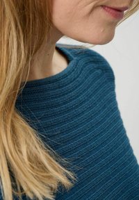 Türkisfarbener Strickpullover mit strukturiertem Rippenmuster, rundem Halsausschnitt und schmaler Passform, kombiniert mit langen, teilweise sichtbaren Haaren.
