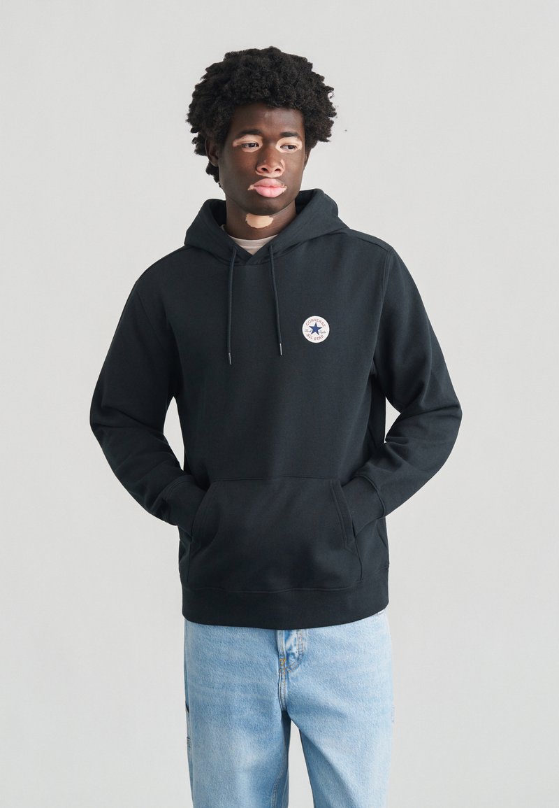 Sort hættetrøje med forlomme, snøre og cirkulært logo på brystet. Bruges sammen med lyseblå jeans.