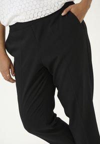 Pantalons noirs ajustés en tissu lisse, dotés d'une taille haute, d'un devant plissé et de poches latérales. Texture douce avec un design épuré.