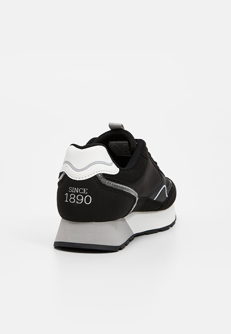 Zapatilla deportiva negra con un panel trasero blanco, acentos grises y tela texturizada. Presenta la inscripción "DESDE 1890" impresa en la parte trasera.