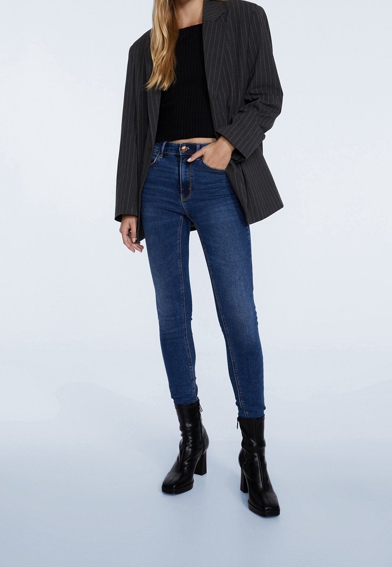 Dark gray pinstripe blazer over a black knit top and blue skinny jeans. Model wearing black high-heeled boots. Smooth fabric, tailored fit.