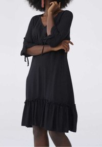 Svart klänning med 3/4 ärm, V-ringning och volangkant. Tillverkad av lättviktsmaterial, med spetsdekoration på ärmarna och justerbara snoddar.