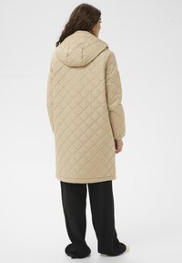 Abrigo beige acolchado con capucha, con un diseño de rombos, mangas largas y un dobladillo de corte recto. Llevado sobre pantalones negros.