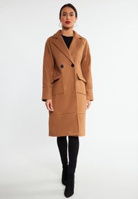 Cappotto lungo in mistura di lana marrone con design a doppio petto, ampie rever, tasche frontali e bottoni neri. Indossato sopra leggings neri.