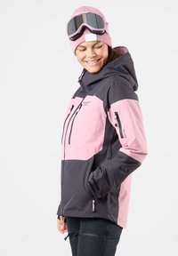 Rosa und schwarze Skijacke mit Kapuze, mit Reißverschlusstaschen und verstellbarem Schnitt. Getragen mit einer passenden rosa Mütze und Skibrille.