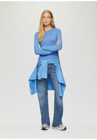 Langärmliges, hellblaues Oberteil mit taillierter Passform, kombiniert mit lockeren blauen Jeans und einem blauen Hemd, das um die Taille gebunden ist; silberne Sneakers.
