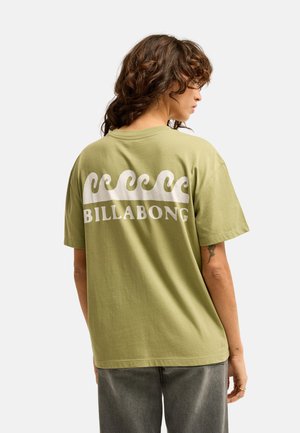 Osoba o kręconych włosach, ubrana w jasnozielony t-shirt Billabong z białym wzorem fali i napisem na plecach, w połączeniu z szarymi spodniami.