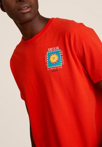 Rode katoenen t-shirt met een vierkant grafisch ontwerp met een zonmotief en een kleurrijke rand. De tekst luidt: “SICILIA IL DOLCE FAR NIENTE LA BELLA VITA.”