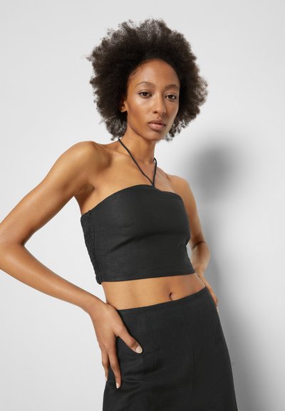 Crop top preto sem alças feito de um tecido texturizado, apresentando um design ajustado e detalhes laterais. Modelo posa com as mãos nos quadris.