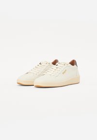 Hvite skinn-sneakers med teksturert overflate, rund tå og tan gummisåle. Med beige snøringer og en brun lærhælakkent.