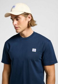 Marinblå bomulls t-shirt med rund hals och korta ärmar, med en vit fyrkantig patch med blå text. Accessoar: ljusbeige basebollkeps.