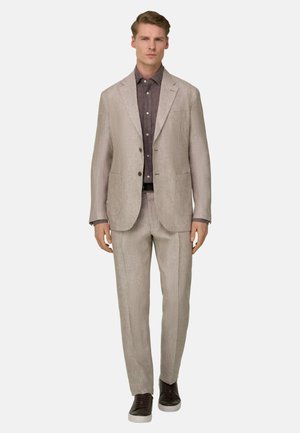 Homme debout portant un costume beige avec une chemise marron foncé boutonnée et des baskets noires sur un fond blanc.