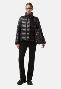 Schwarze Daunenjacke mit hohem Kragen und gestepptem Design, kombiniert mit schwarzen Hosen und einer passenden gesteppten Tasche, dazu klobige schwarze Schuhe.