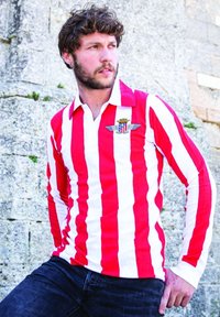 Rood-witte gestreepte lange mouwen shirt met een kraag en een logo met vleugels en een kroon. Gemaakt met donkere spijkerbroeken tegen een stenen achtergrond.