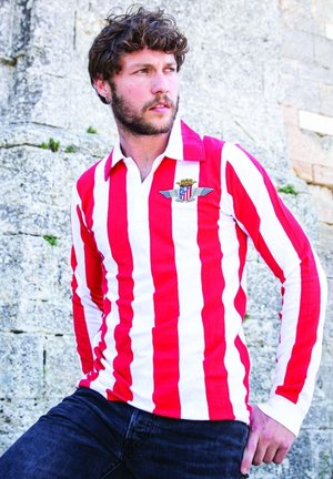 Rood-witte gestreepte lange mouwen shirt met een kraag en een logo met vleugels en een kroon. Gemaakt met donkere spijkerbroeken tegen een stenen achtergrond.