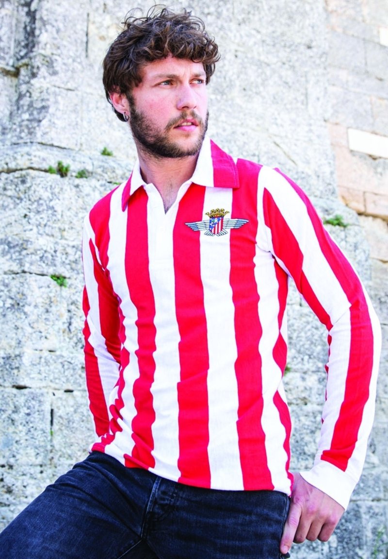 Rood-witte gestreepte lange mouwen shirt met een kraag en een logo met vleugels en een kroon. Gemaakt met donkere spijkerbroeken tegen een stenen achtergrond.