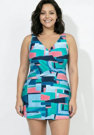 Uśmiechnięta kobieta z ciemnymi włosami do ramion, ubrana w bezrękawnikową sukienkę w wielokolorowy geometryczny wzór, na tle jednolitej białej ściany.