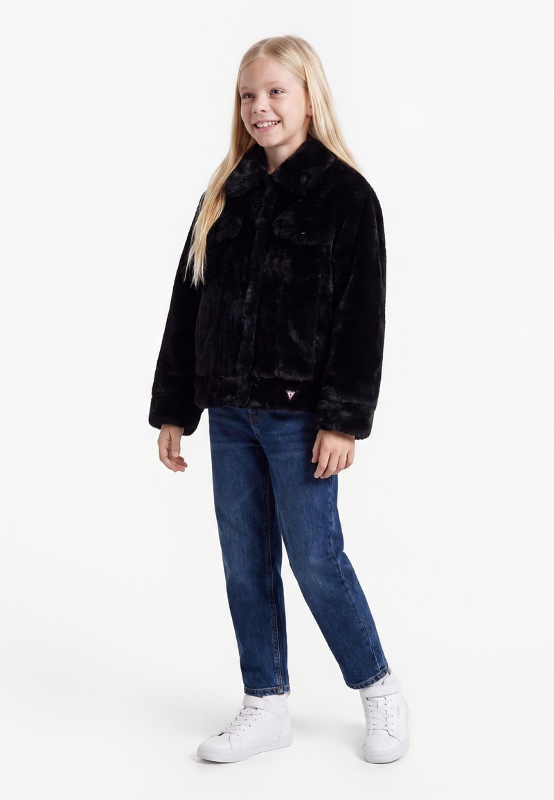 Jeune fille souriante aux longs cheveux blonds, vêtue d'une veste en fausse fourrure noire, d'un jean bleu et de baskets blanches, debout devant un fond blanc uni.