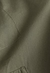 Tissu vert olive avec une texture subtile, présentant des coutures visibles et une zone de superposition de tissu. Aucun motif distinct ni élément métallique.