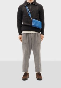 Maglione lavorato a maglia scuro con colletto zip, pantaloni di velluto a coste grigi, scarpe stringate marroni e una borsa a tracolla in pelle blu.