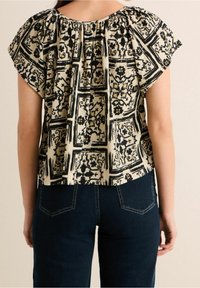 Blusa a maniche corte con motivo nero e crema, caratterizzata da una vestibilità ampia e un colletto arricciato. Indossata con jeans blu scuro.