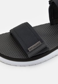 Sandalia negra con suela de goma texturizada, correas de tela y marca Columbia. Presenta una entresuela blanca lisa y un patrón angular en la suela.