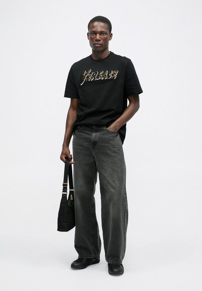 T-shirt preta de algodão com texto em estampado metálico de leopardo, combinada com jeans de perna larga cinza-escuro. Uma bolsa preta com acabamento texturizado está segurada.