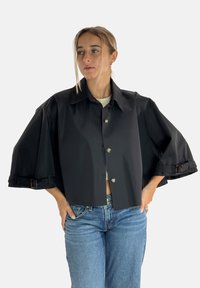 Schwarze kurze Jacke mit weiten Ärmeln, geknöpftem Vorderteil und verstellbaren Riemen an den Handgelenken. Glatter Stoff und strukturierter Kragen.