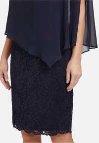 Femme portant une jupe crayon en dentelle bleu marine avec ourlet festonné et un haut overlay transparent bleu marine avec ourlet asymétrique et fente au bras.