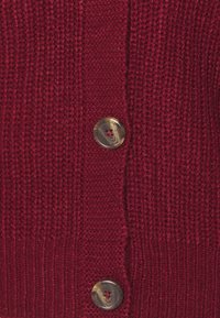 Gebreid vest in diep rood met geribbelde textuur. Voorzien van ronde, bruine knopen met een subtiele glans en contrasterende stikseldetails.