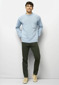 Junger Mann steht lächelnd mit Händen hinter dem Rücken, trägt einen hellblauen Strickpullover, dunkelgrüne Hose und beigen Sneakern vor einem weißen Hintergrund.