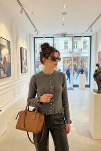 Femme avec lunettes de soleil, pull gris boutonné et sac à main marron se tient dans une galerie d'art moderne lumineuse avec des peintures et des sculptures.