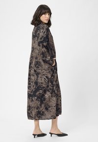 Rochie lungă cu un model spiralat în nuanțe de negru și brown, fabricată dintr-un material moale. Prezintă o croială lejeră, mâneci lungi și buzunare laterale, purtată cu mule negre.