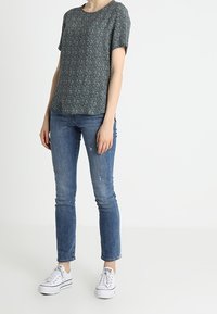 Kvinde iført en kortærmet mørk blomsterbluse, tætsiddende blå jeans og hvide sneakers, stående foran en hvid baggrund.